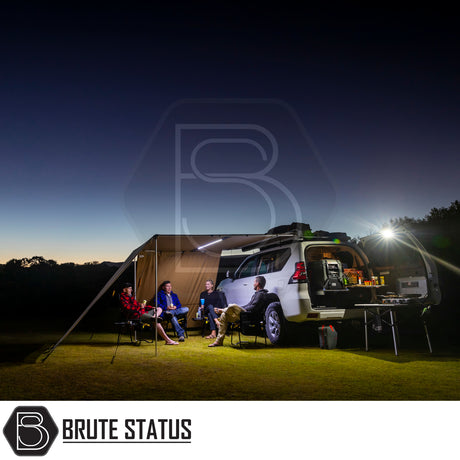 White SUV with ARB PVC Case Awning & LED Light Kit extended; people seated under awning, camp kitchen illuminated by LED lights, set in a grassy twilight clearing.