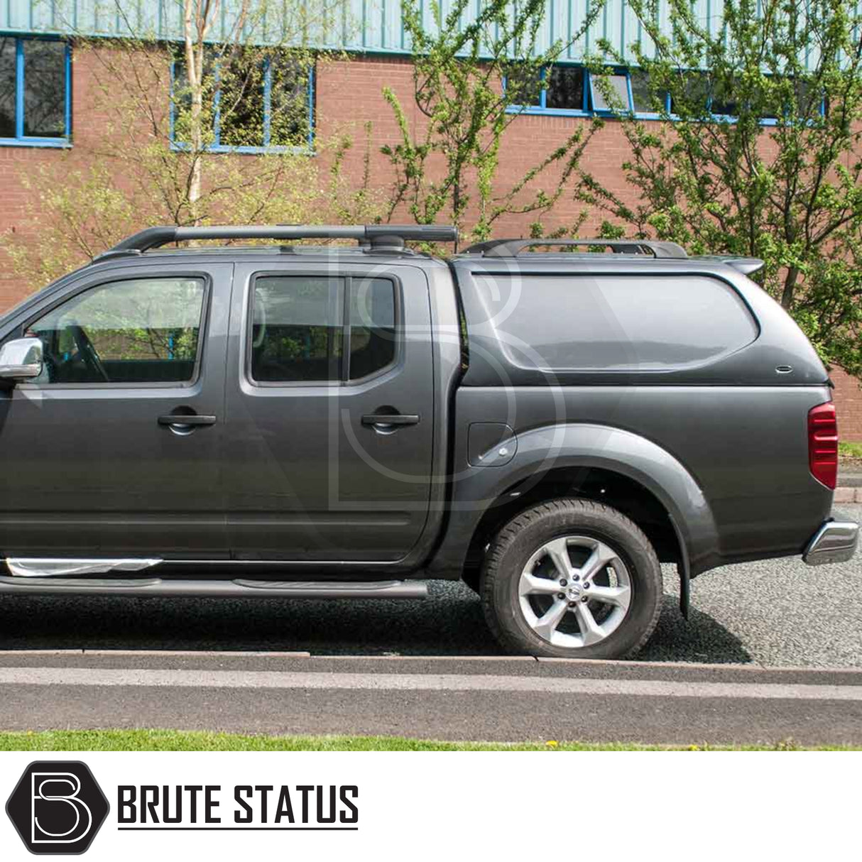 Grey Nissan Navara 2005-2015 D40 with Truckman L-Series Hardtop Canopy, featuring alloy wheels, roof rails, and secure remote locking, parked by a brick building.