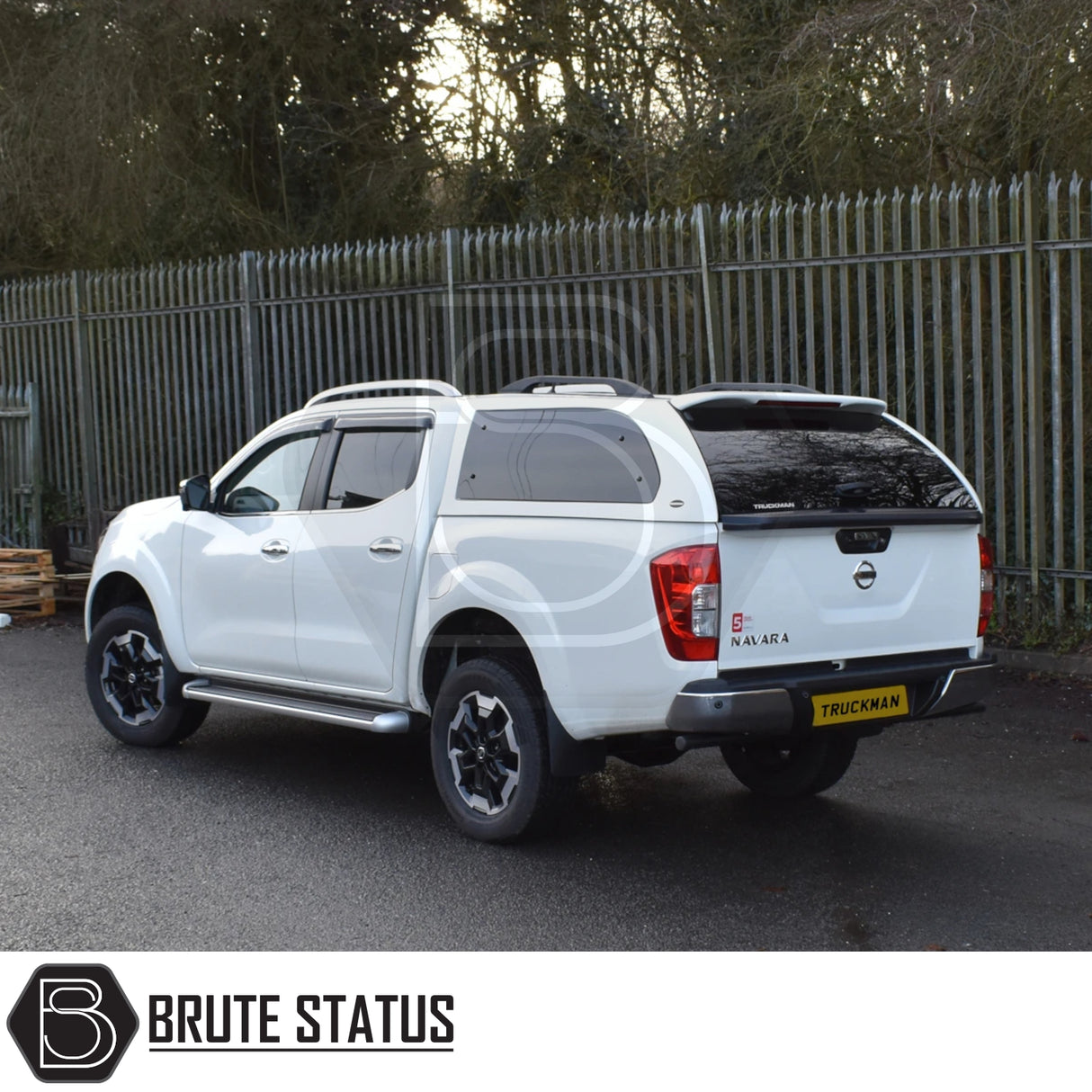 Nissan Navara pickup with Truckman Grand hardtop canopy, featuring remote locking and tinted windows, parked on wet asphalt beside a metal fence.