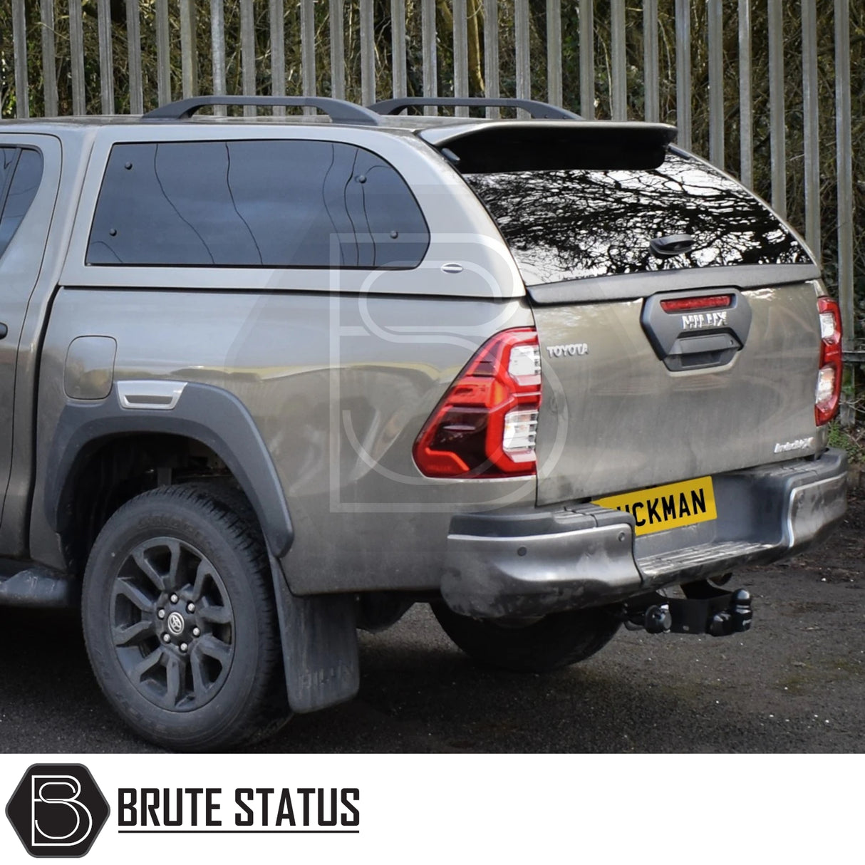 Rear view of grey Toyota Hilux Invincible X with Truckman Grand Hardtop Canopy, featuring tinted windows, roof rails, tow hitch, and Brute Status watermark.