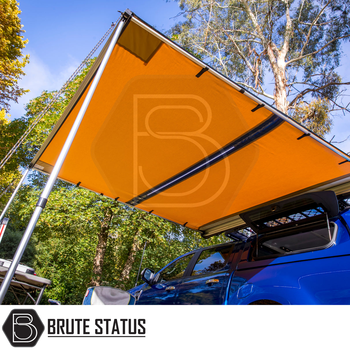 Orange vehicle-mounted awning from Brute Status, extended from a blue pickup, creating shaded camping space with table and chair; ARB Flinders Rooftop Tent context.