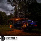 Dusk camping scene with SUV featuring ARB Aluminium Awning & LED Light Kit, folding chair, table with stove, and open rear storage.