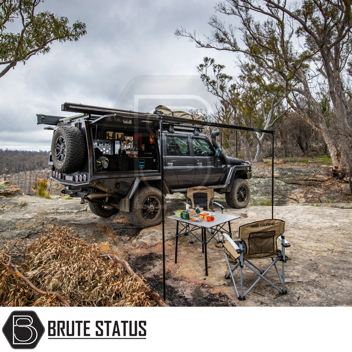 Off-road 4x4 setup with ARB Flinders Rooftop Tent, canopy, awning, gear shelves, and camping chairs, showcasing rugged self-sufficient camping on a rocky ledge.