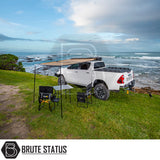 Toyota truck with ARB Aluminium Case Black Awning & LED Light Kit, camping setup beneath, overlooking rocky shoreline, featuring retractable awning and chairs.