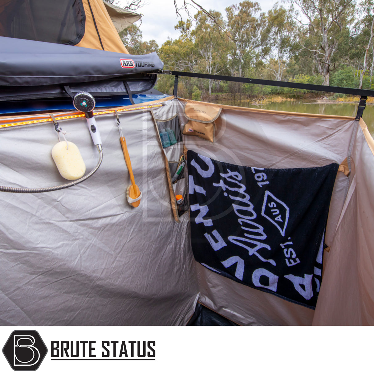 ARB Ensuite Room Shower Tent set up in a bush-camping environment, featuring a handheld shower, toiletries organizer, and towel, highlighting privacy and convenience in outdoor settings.
