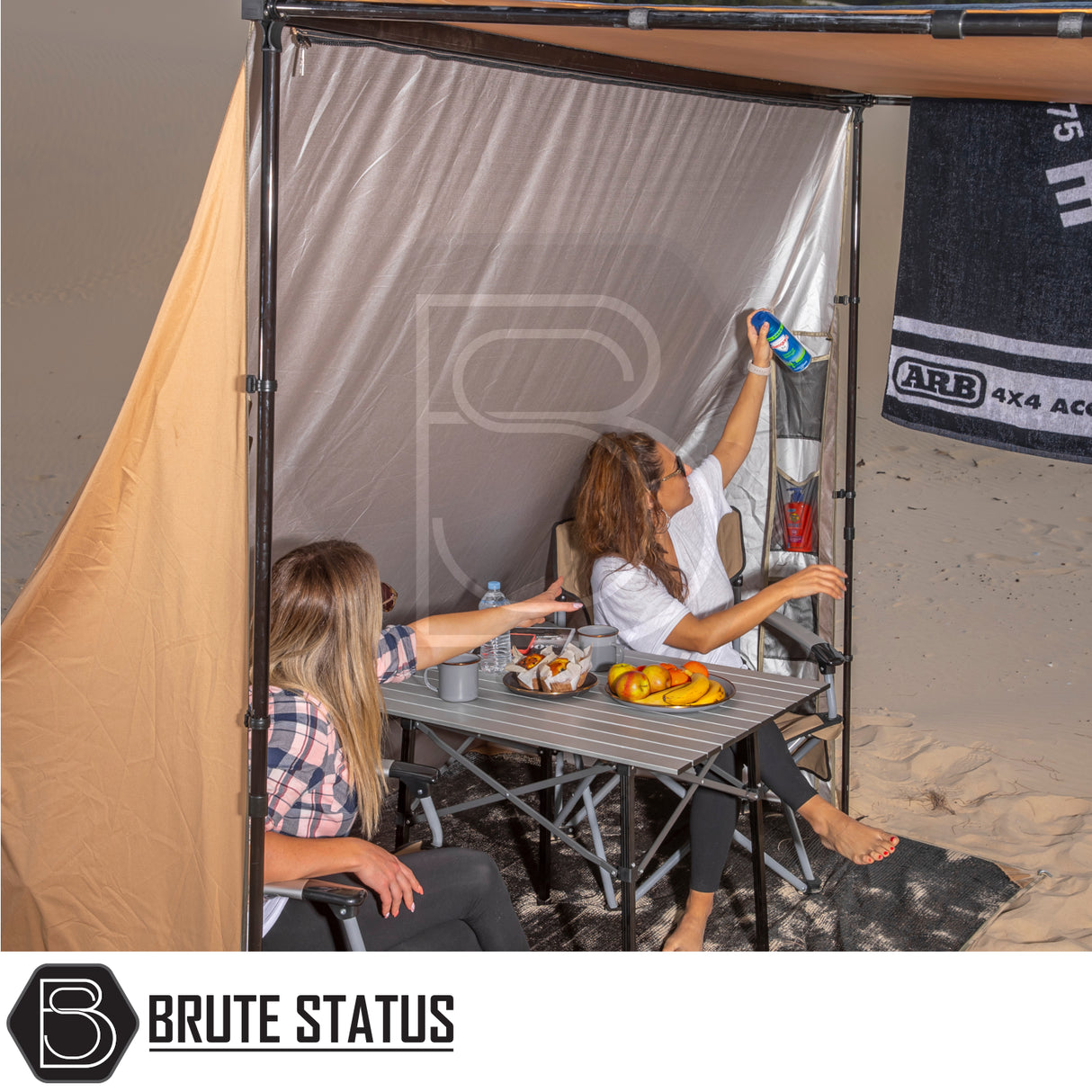 Two women enjoy a beach campsite under a Brute Status-branded ARB Awning Alcove, showcasing its versatile protection and organized storage features.