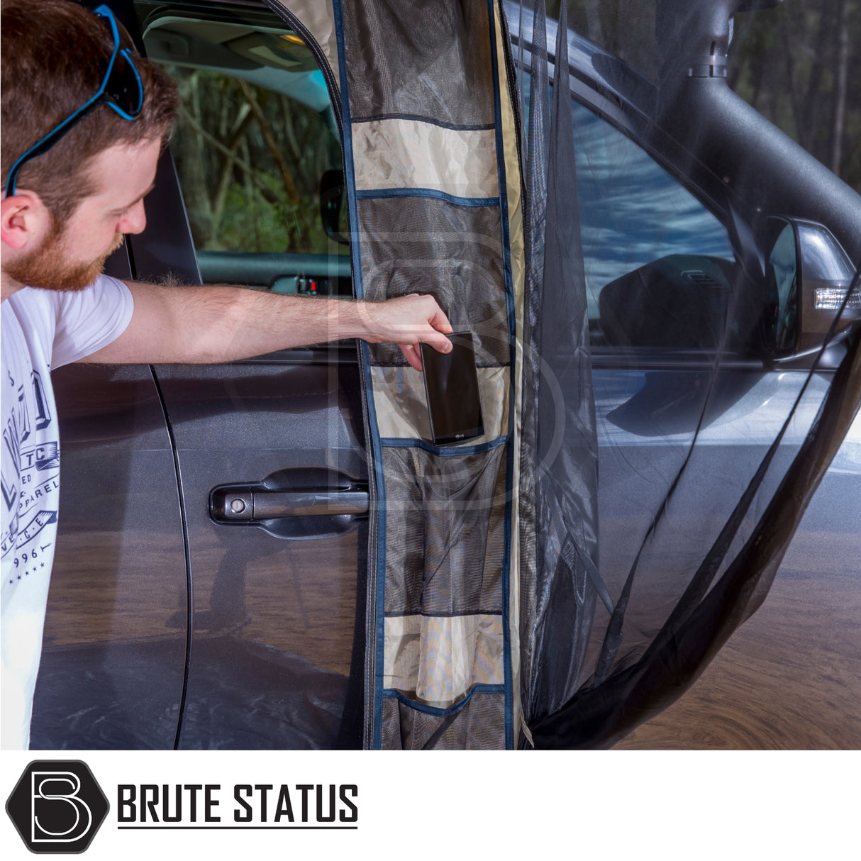 Man placing phone in mesh pocket of Brute Status SUV bug-net curtain, part of ARB PVC Case Awning & LED Light Kit, attached to open vehicle door.