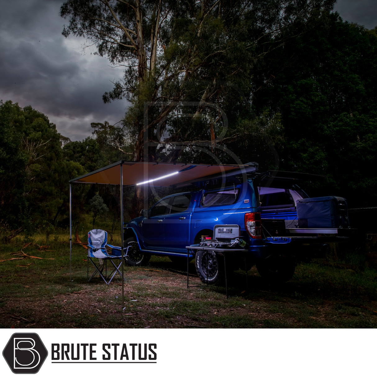 ARB Flinders Rooftop Tent on blue 4x4 ute, showcasing camping setup with awning, camp kitchen, and storage in wooded dusk setting by Brute Status.