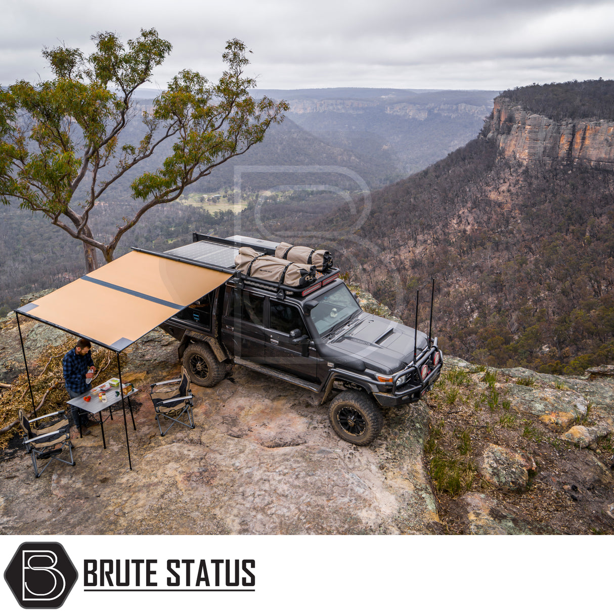 Off-road vehicle on cliff with ARB Aluminium Case Awning & LED Light Kit extended, offering shade. Camping setup includes table, chairs, and person preparing food.