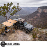 Off-road vehicle on cliff with ARB Aluminium Case Awning & LED Light Kit extended, offering shade. Camping setup includes table, chairs, and person preparing food.