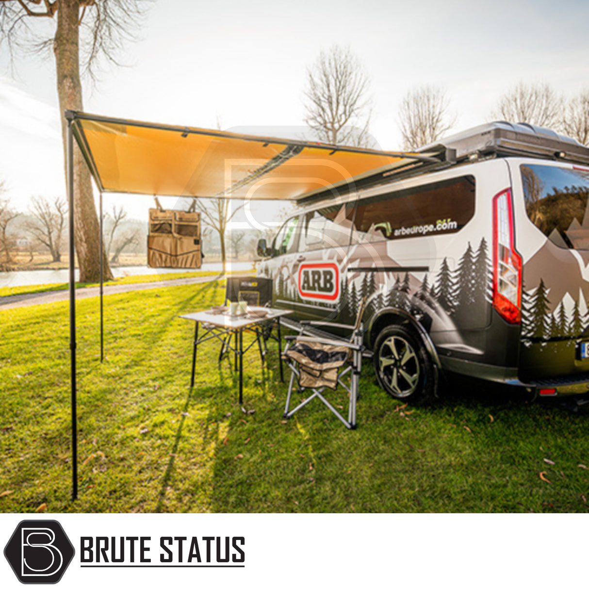 Overland-style camper van with ARB Awning with Light Black Aluminium Case 3m x 2.5m, featuring an awning, folding table, and camp chair by a riverside.