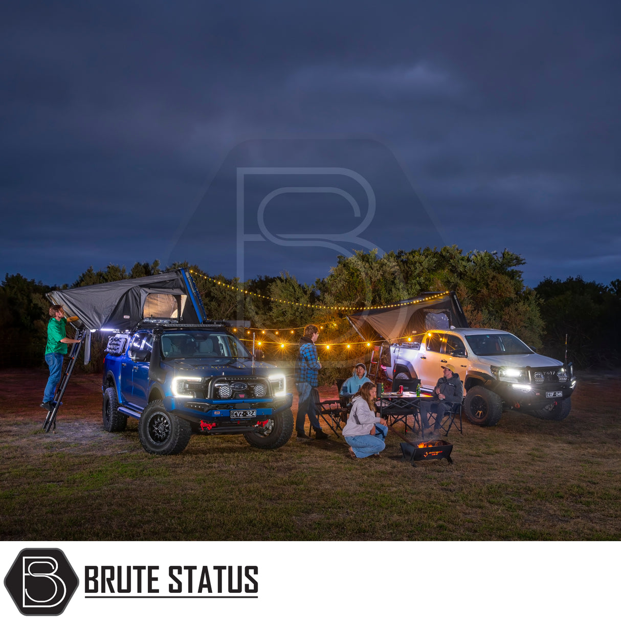 Camping scene with two pickup trucks, each fitted with ARB Esperance 2.0 Rooftop Tent Hard Shells, string lights, and a portable fire pit.