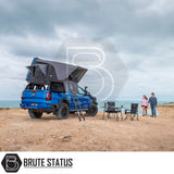 Rooftop tent on a Ford Ranger at a beach, showcasing the ARB Esperance 2.0 Rooftop Tent's setup with ladder and camping gear, under Brute Status branding.