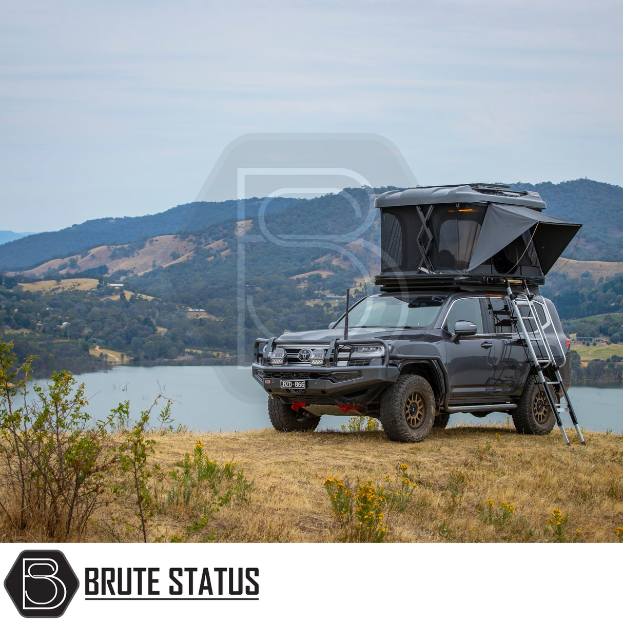 Toyota Land Cruiser-style SUV with ARB Altitude Rooftop Tent, featuring a pop-up design and ladder, parked on a grassy bluff overlooking scenic lake and hills.