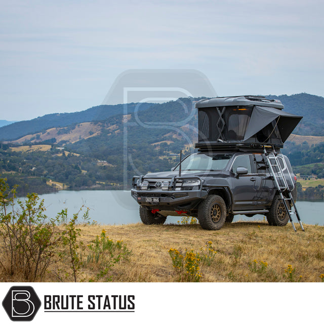 Toyota Land Cruiser-style SUV with ARB Altitude Rooftop Tent, featuring a pop-up design and ladder, parked on a grassy bluff overlooking scenic lake and hills.