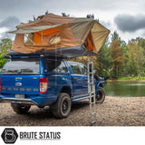 Blue Ford Ranger with ARB Flinders Rooftop Tent on rocky riverbank; fold-out design, brown canvas, aluminum ladder, ideal for adventurous camping.