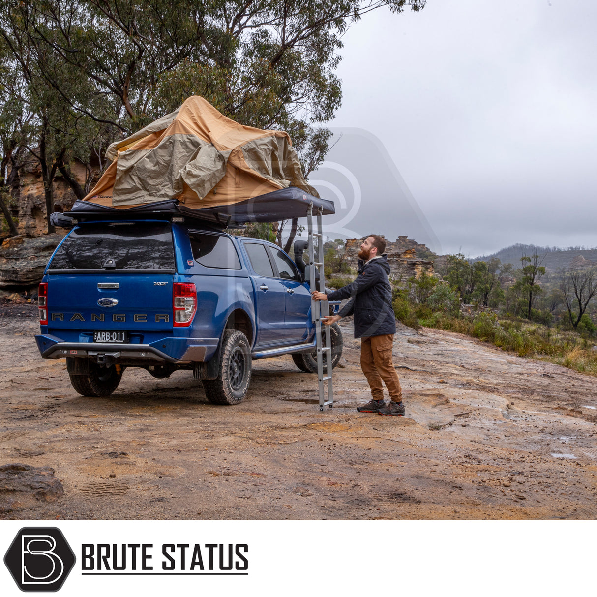 Blue Ford Ranger with ARB Flinders rooftop tent setup, man adjusting ladder in bushland; showcases Brute Status's premium off-road camping gear.