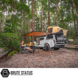 Overland pickup with ARB Flinders Rooftop Tent, slide-out kitchen, and canopy at a forest campsite. Two people relax under an awning.