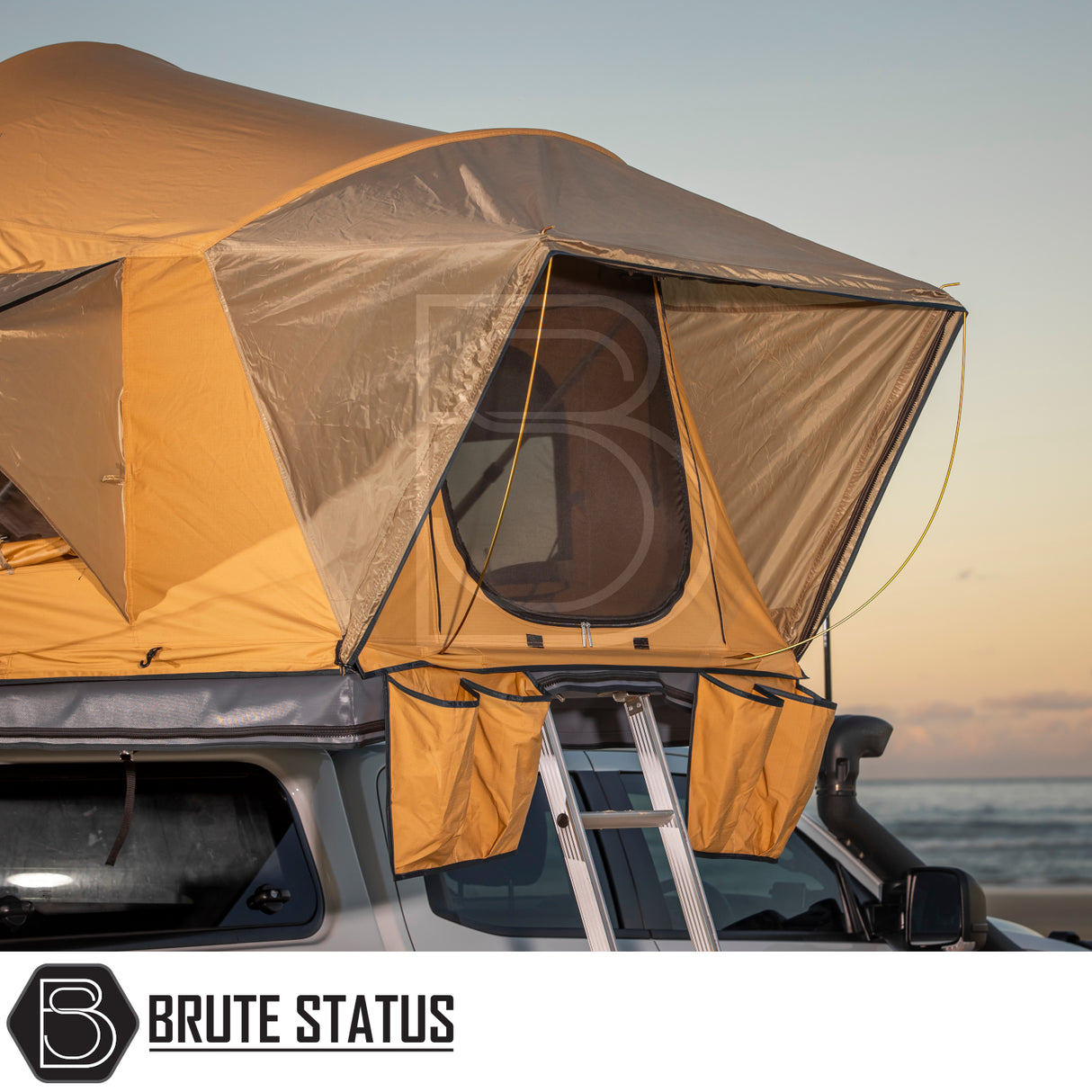 ARB Flinders Rooftop Tent on a vehicle, mustard-tan canvas with mesh windows, aluminum ladder, and storage pockets, set against a sunset by the sea.