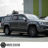 Grey VW Amarok with Truckman L-Series Hardtop Canopy, showcasing aftermarket design, chrome side-steps, alloy wheels, parked by industrial building.
